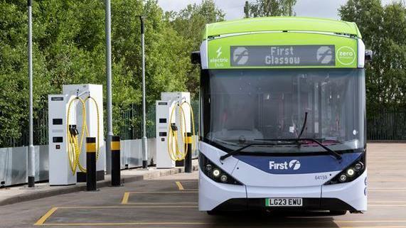 Single decker bus, white and blue at the front with "First" written below the front window. The roof is lime green with the destination board at the front reading "First Glasgow". The bus is parked in an outdoor carpark area, next to two large white electric vehicle chargers with yellow cables.