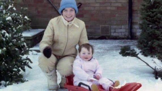 A baby in a pink all-in-one snowsuit, sitting on a red plastic sledge. A woman in a beige snowsuit and light blue beanie hat squats down next to her. Both are smiling at the camera.