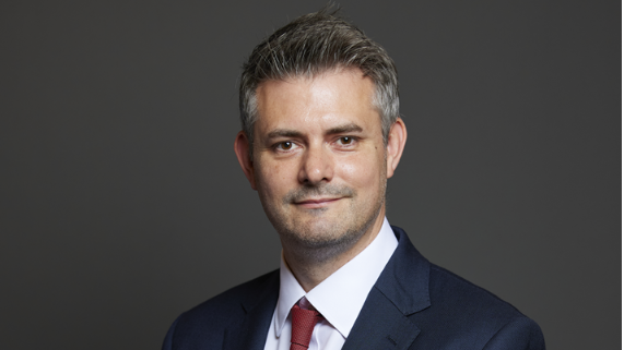 Man with short grey hair blue blazer white shirt red tie infront of grey background, official portrait 