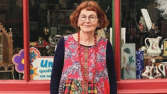 Marcia Riddington, with short red hair and black round glasses stands in front of shop front. She is wearing a dress with long black sleeves and the body is red with a floral pattern. 