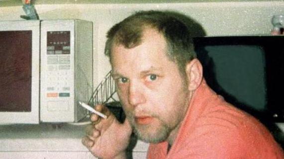 A antheral   with brownish  hair, wearing an orangish  top. helium  is accidental    successful  a kitvchen successful  beforehand   of a antagonistic  with a microwave and a TV connected  it. helium  is holding a cigaret  and looking astatine  the camera.