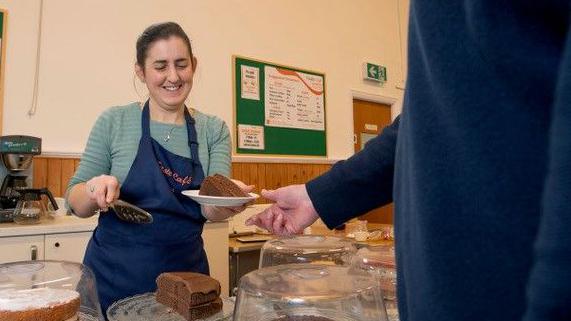 A woman behind a cake counter at Ebenezer Church in Horfield in Bristol serves a man with a slice of cake. She is smiling and wearing a blue apron