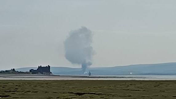 A large cloud of smoke rises above the scene. Ancient ruins can be seen on the left by an expanse of water and grass in the foreground. Hills form the backdrop.