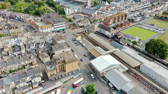 The image shows an aerial view of a busy urban area and a train station. The station has multiple platforms and tracks, with several trains visible. Nearby, there’s a bus terminal with several buses.