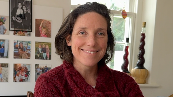 A woman in her late thirties sat in front of an electric piano wearing a burgundy knitted jumper. she has her hair in a short bob and is smiling 