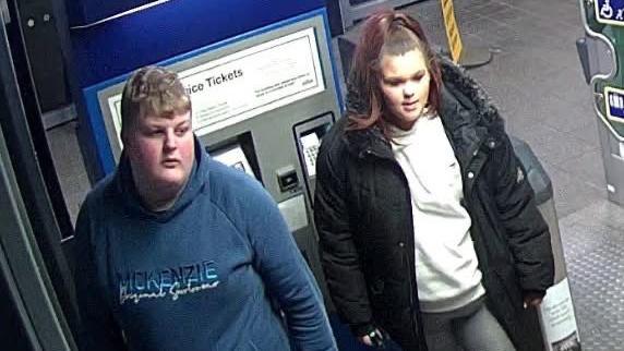 Two teenagers captured on CCTV at a train station: a boy in a navy McKenzie tracksuit with blond hair on the left and a girl in grey leggings and a black fur-hooded coat with her hair tied up on the right. Both are facing away from the camera.