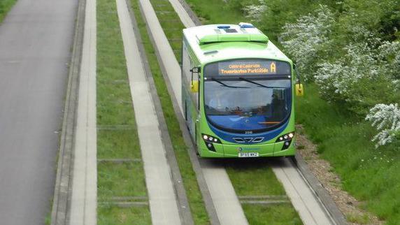 A green single-decker busway bus pictured at Over in Cambridgeshire. The busway has two lanes and has a grassed area between the concrete runners for the bus wheels. A tarmac footpath runs parallel to the busway on the left with bushes on the right of the busway.