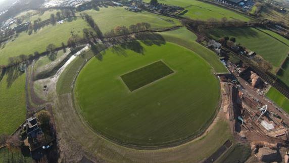 An aerial view of a circular grass pitch with a construction site to the bottom left 