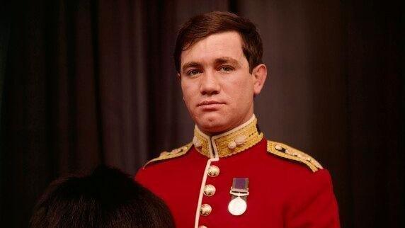 A cropped version of a National Portrait Gallery image of the Army officer Captain Robert Nairac. He has short, brown hair and is wearing a red military jacket decorated with gold buttons, gold epaulettes and single medal pinned to his chest. He is holding a tall, black bearskin hat.