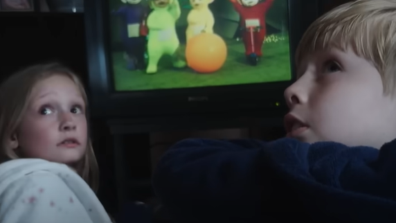 Two child actors looking scared with the Teletubbies on the TV in the background.