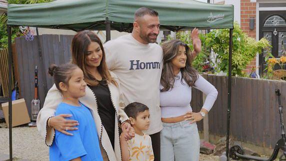 Sheeny and her husband Sohan standing next to their three children in front of a green gazebo and a wooden fence in a garden.