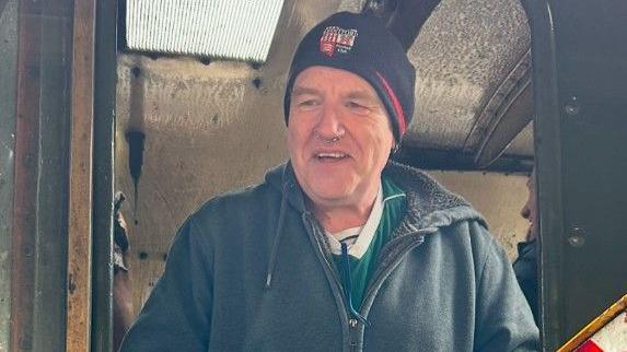 Martin Sullivan, smiling, wearing a dark hat, dark hoodie and top, and standing in the doorway of an old train carriage. 