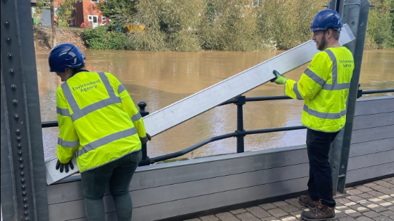 Two men in yellow high visibility jackets liftin a metal beam into place between two metal uprights, forming a wall with the river in the other side