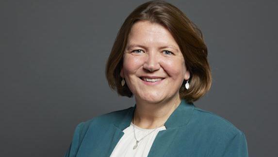 A woman wearing a green/turquoise jacket with a white top looks towards the camera and smiles 