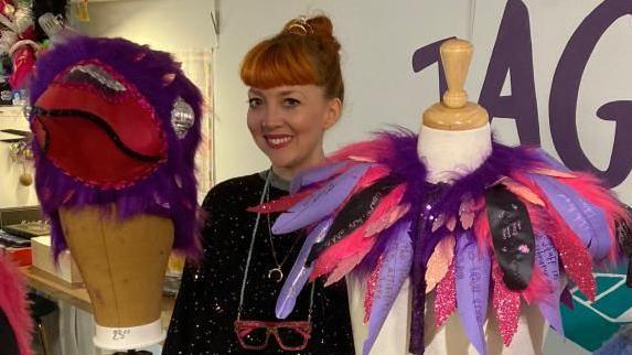 A fashion designer stands in the middle of the two pieces of the mascot on mannequins. On one side is the crow's head, which is bright purple and read and fluffy. On the other side is a neck pece made up of black, pink and purple feathers with messages on. The designer has red hair, which is tied up, and is wearing a black, sparkly top. A pair of red glasses hang on a chain from her neck and she is smiling.