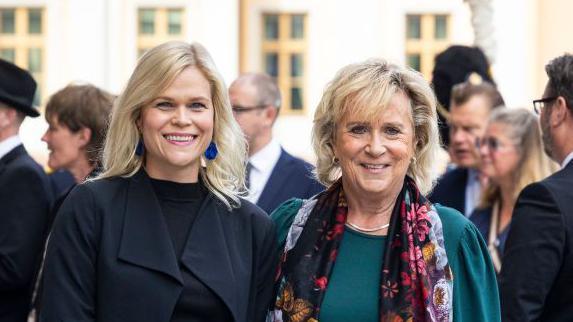 Headshot photo of Swedish minister Paulina Brandberg (left) and another unidentified person on the right. Brandberg is dressed in black clothes and a black coat and is smiling.