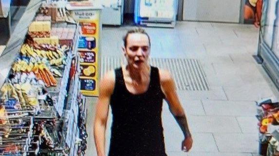 CCTV still of Rachel Booth with blonde hair tied back tightly. She walks past shelves of snacks to the garage till. She is wearing a sleveless black top.