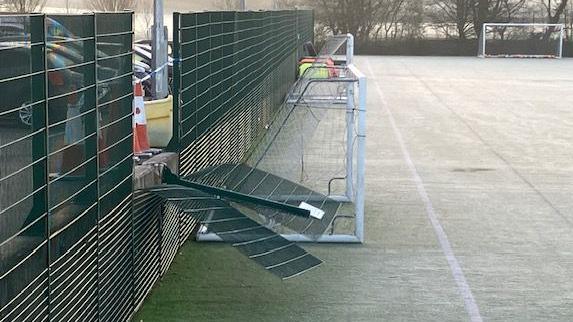 A section of the green fence around the football pitch has been bent at a 90 degree angle. Orange traffic cones are lined in front of the gap while blue and white police tape cordons off the scene. On the pitch side of the fence are two five-a-side goals lined up along what would be the touchline of a normal size pitch. A full size goal can be seen in the distance.