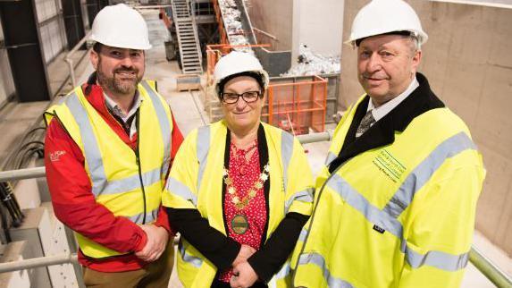 Council leader Kevin Guy next to cabinet members Sarah Moore and Tim Ball. All three in hi viz vests, standing on a balcony with the new sorting area for household recycling in the background.