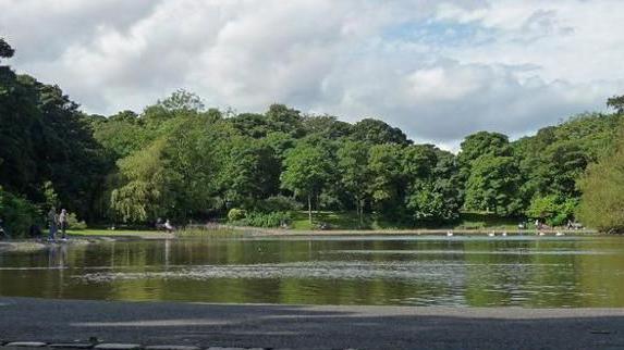 The lake at Leazes Park in Newcastle dominates the image with trees around it. A number of people are standing or walking around its edge.