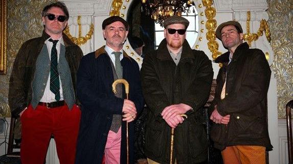 The Beta Band look into the camera - two of the band members are holding canes, while three have flat caps. Two have glasses or sunglasses on. They are standing in front of regal surroundings, including a large mirror and a white and gold mantelpiece 