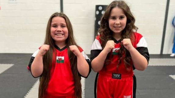 Lili-Rose and Annarose face the camera in their Team Wales tracksuits, smiling and with arms raised 