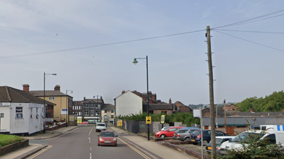 A road with an orange car and white van travelling down the right-hand carriageway. A car park on the far right of the picture and a junction on the left-hand side next to a white building. 