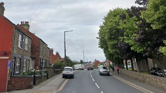 A road with terraced houses on one side and a row of tress on the other side. Pedestrians walking on the pavement.