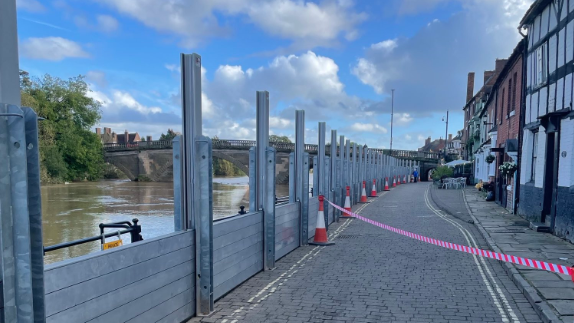 A road with houses running down one side and metal posts on the other, with slats running between them to form a wall, with the river on the other side and a bridge in the distance