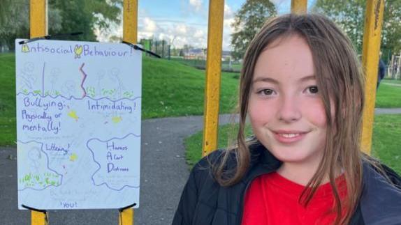 10-year-old Mia with brown hair, some of it down some of it tied up smiling into the camera, wearing a black coat and red jumper. She is standing next to her anti-social behaviour sign which is tied to bars in the park.