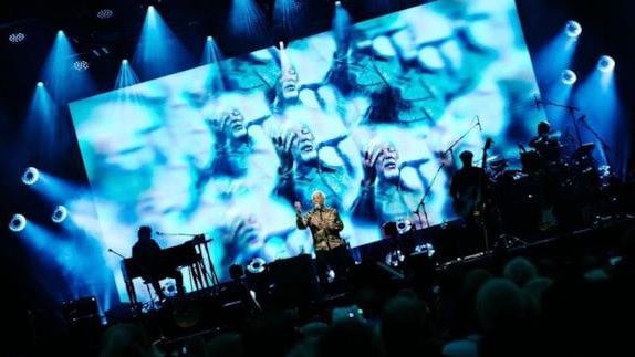 Sir Jones sings on stage while his  band, performing on piano and drums, are silhouetted by a large blue screen.