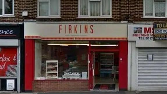 A bakery with "FIRKINS" on its signage on the high street with red columns either side of it stands next to a shopfront with metal shutters down with a sign reading "TO LET" above it.