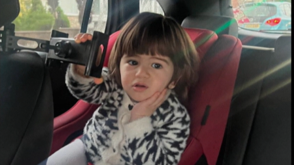 Toddler Mila sitting in a car seat holding her face and a bracket similar to an extendable TV wall mounting. She wears a black and white woolly cardigan.