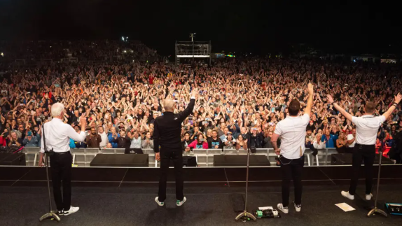 Four men standing on a stage with their backs to the camera waving to a large crowd facing them