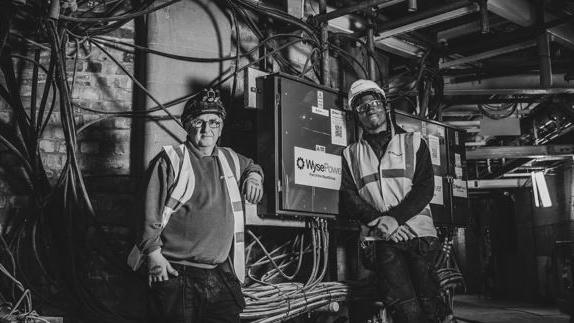 Two tradesmen in hard hats lean against a cabling box in the town hall.