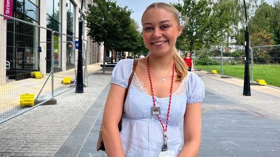A blonde-haired woman smiling wearing a red lanyard and white short-sleeved blouse.