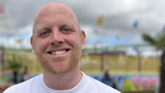 Man smiles and looks at camera. Behind him is a fairground attraction.