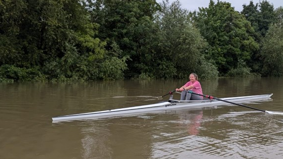 Julie in action in a single scull rowing boat on a river with trees in the background 