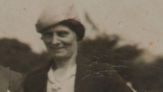 Black and white image of Louisa Dunne. It is an old and soft focused photo. She has dark hair tied back and is wearing a light-coloured hat and dark coat.