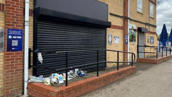 The entrance to the former club shop, which has a small ramp leading to it. The entrance is covered by black shutters. There are a number of flowers left outside of it.