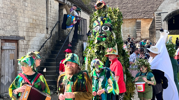 A person in a large green leaf coloured costume makes it way past an old brick building as people dressed in green with painted faces walk nearby.