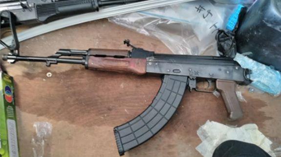 An AK-47 is positioned on a wooden board. There is plastic wrapping above the rifle which is made of black plastic, metal and wood. 