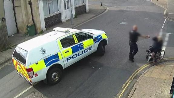 A police truck parked in the middle of King Street, Great Yarmouth, with the police officer, arm extended, deploying PAVA spray on a man, who is in his wheelchair on the white line of the junction of St Peter's Road. Behind the police van is the Old White Lion pub, which is converted into flats, and to the right of the image is the frontage of the Londis convenience store. In the background is King Street looking towards Great Yarmouth town centre.