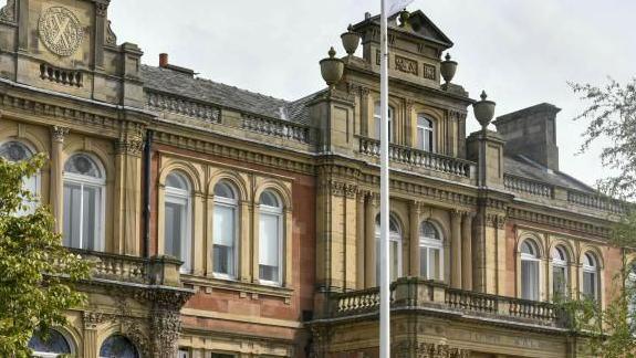 Frontage of Penrith town hall, a late 18th Century sandstone and red brick building with ornate lintels and balconies and decorative stone urns on either side of the upper windows. A few trees are just poking into shot. A white flag pole stands in front.