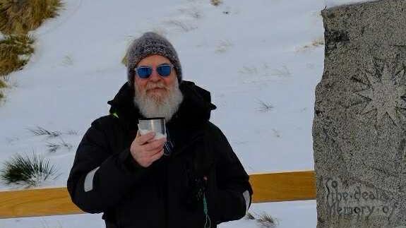 Steve Winn holds up a flask cup while standing next to a memorial stone. He is surrounded by snow and is wearing a black winter jacket and a grey woolly hat. He has a large full grey beard and is wearing blue tinted sunglasses.