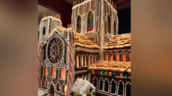 This image shows a detailed gingerbread cathedral, photographed up close. A large rose window made from melted candy sits on the left façade, surrounded by intricate white icing that mimics carved stone tracery. The structure features tall Gothic-style windows, each decorated with vertical lines of icing and filled with stained‑glass–like candy.
