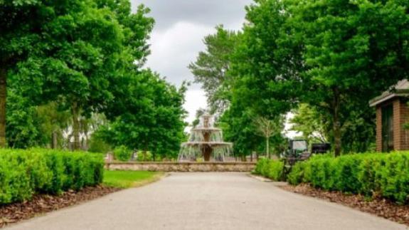Landscaped park with a central fountain surrounded by grass. There are bushes, paving and trees.