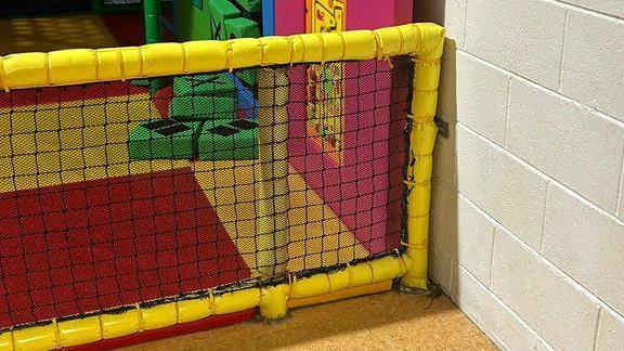 A children's softplay park is surrounded by yellow, foam covered posts and has colourful flooring. On the outside corner, a small brown rat can be seen lying on the floor between a post and the wall. 