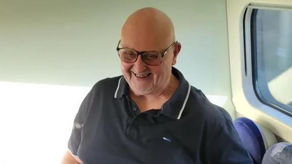 Barry smiles wearing a black polo shirt while sat on public transport, with sun shining through a window. 