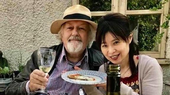 Glyndwr Meredith (left) and his wife hold up a glass of wine and a plate with a slice of pizza on it while posing for a photo. They are next to a table that has a cheeseboard and a bottle of wine in the foreground.